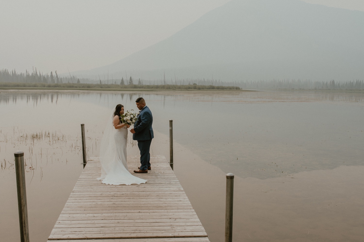 bride and groom standing on dock after first look for a mountain elopement during wildfire season in Banff Alberta