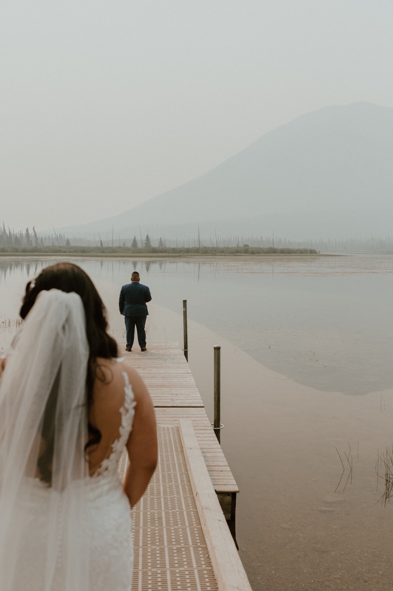 bride walking behind groom on dock to do their first look for a mountain elopement during wildfire season in Banff Alberta