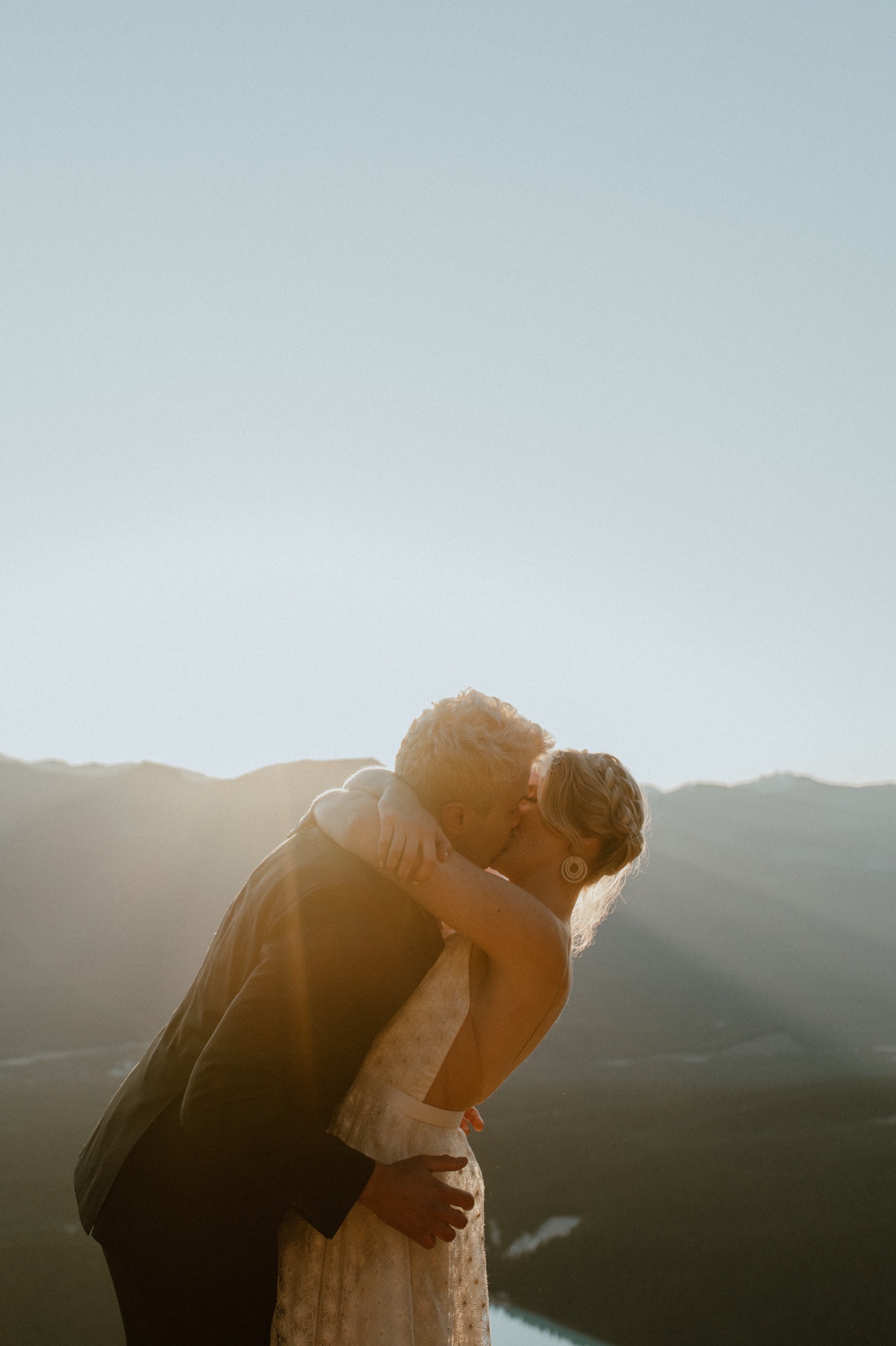 big embraces as the couple kissing and having an intimate moment as the sun rises above the mountain ridge seen from the top fo a mountain by Lake Louise in Alberta