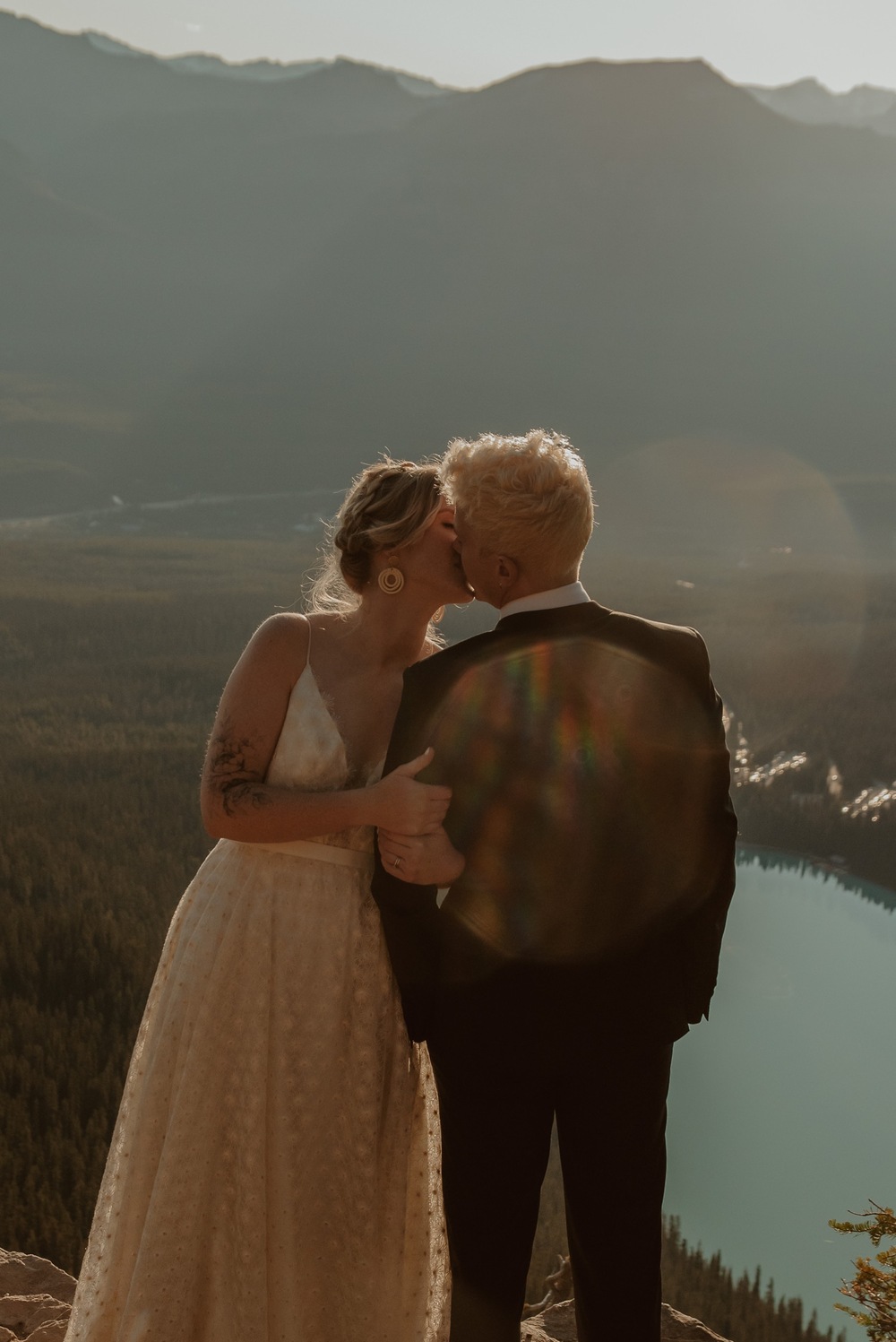 couple embracing and having an intimate moment as the sun rises above the mountain ridge seen from the top fo a mountain by Lake Louise in Alberta