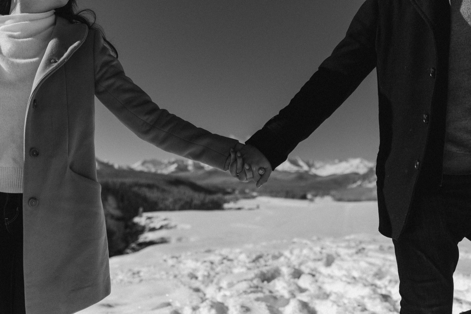 black and white shot of couple holding hands in front of amazing mountain backdrop during a winter engagement session in the rocky mountains of Alberta