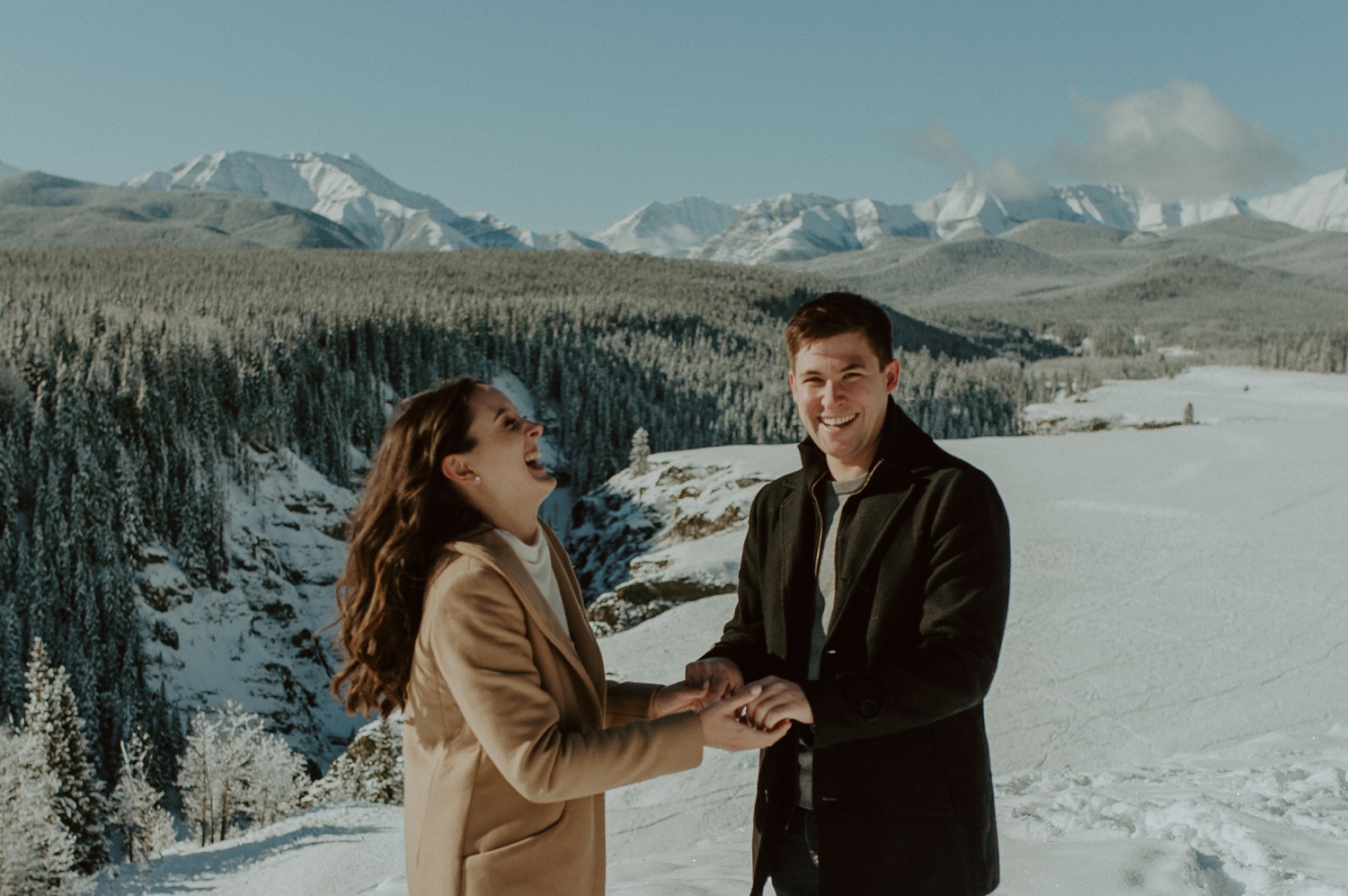 couple laughing candidly to each other in front of amazing mountain backdrop during a winter engagement session in the rocky mountains of Alberta