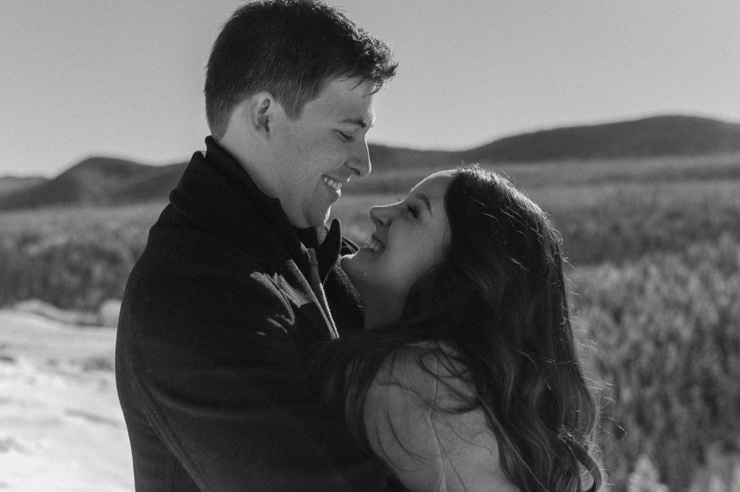 black and white candid shot of couple cuddling in front of amazing mountain backdrop during a winter engagement session in the rocky mountains of Alberta