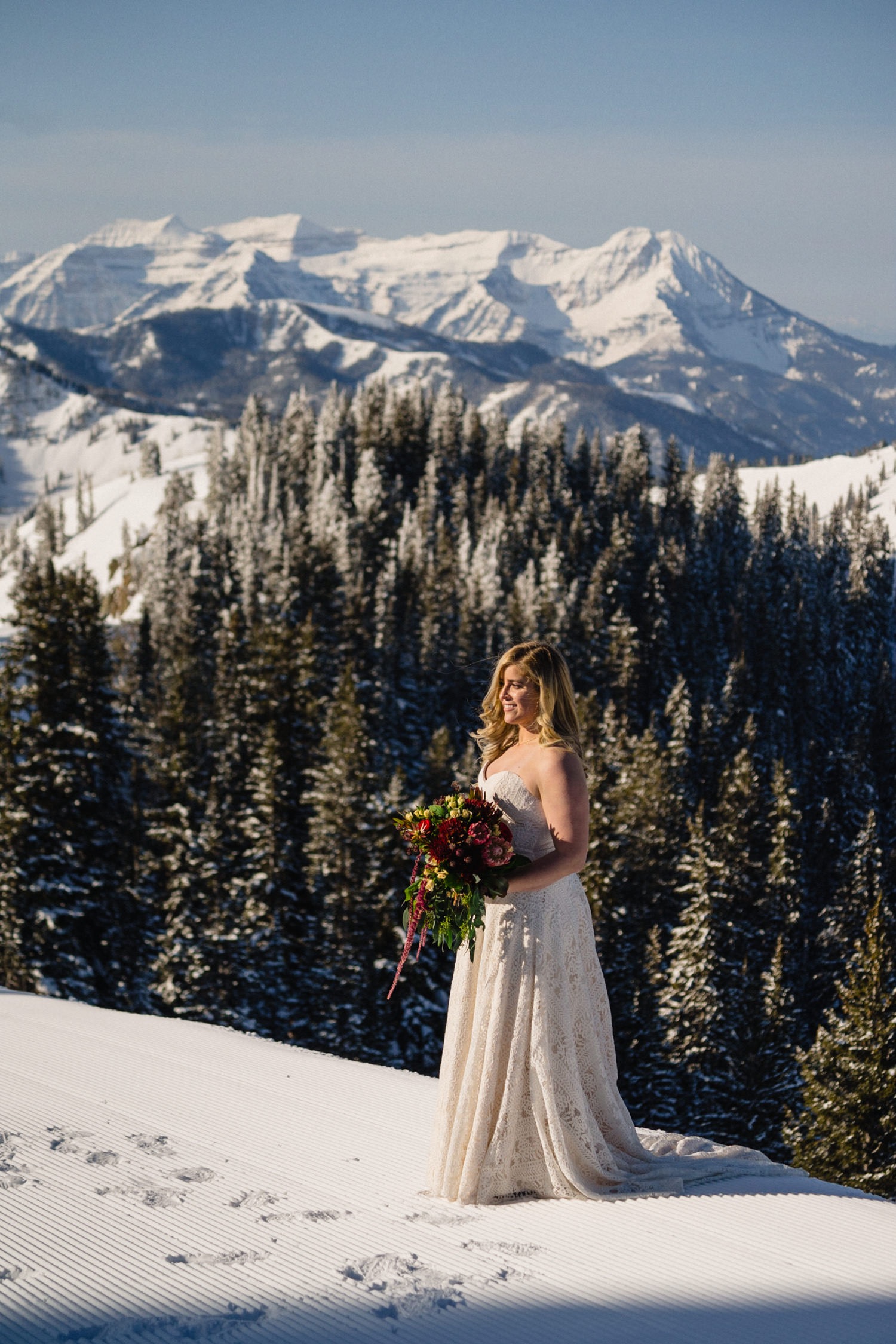 Bride in wedding dress on snowy peak with snow covered mountains behind her. 