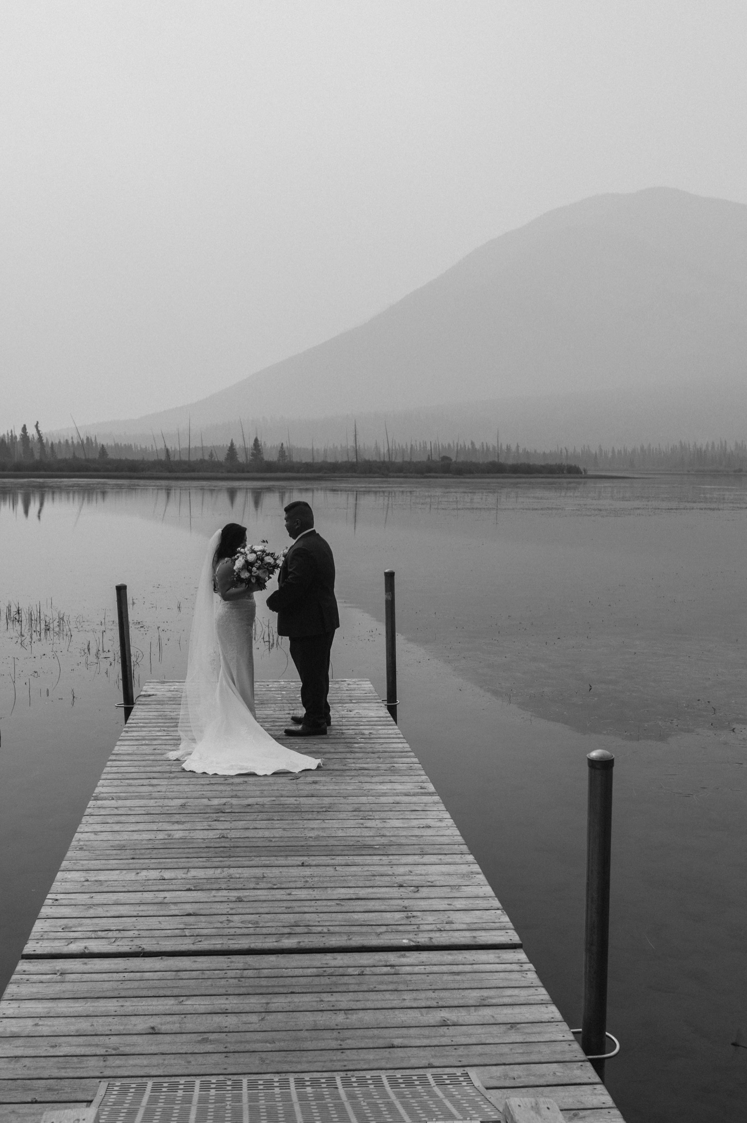 black and white shot of bride and groom standing on dock after first look for a mountain elopement during wildfire season in Banff Alberta