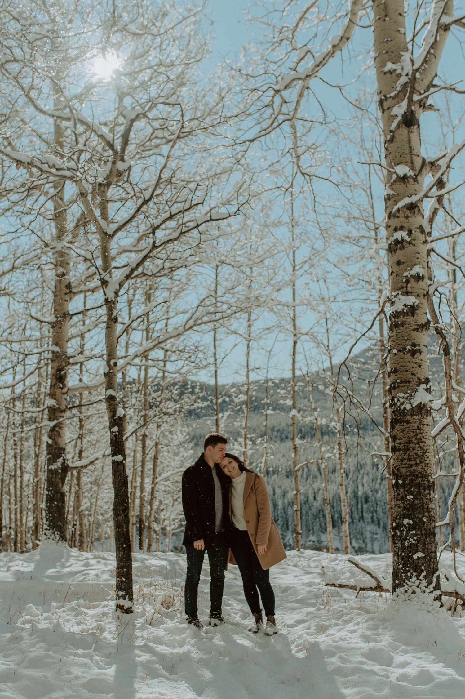 wide shot of couple in forest in front of amazing mountain backdrop during a winter engagement session in the rocky mountains of Alberta